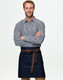 A man in a checkered shirt wears a denim half apron with a brown tie belt. The apron features pockets and a logo.
