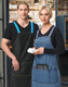 Two models wearing denim aprons with changeable blue and striped straps, featuring pockets for accessories.
