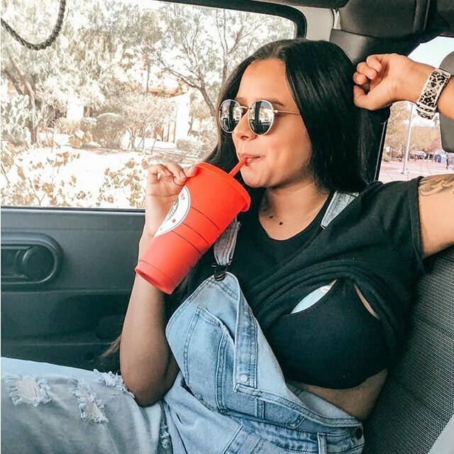 A woman wearing sunglasses and dungarees sits in a car drinking from a red cup with a straw. Her shirt is lifted to show her Elvie Pump tucked inside her bra. 