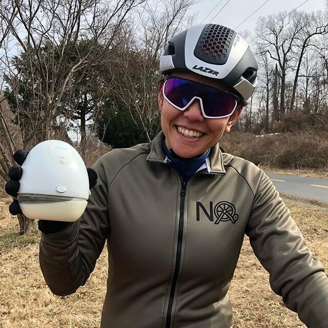 A sportswoman wearing a helmet, sun visor, and sportswear holding up an Elvie Pump filled with breastmilk and smiling. 