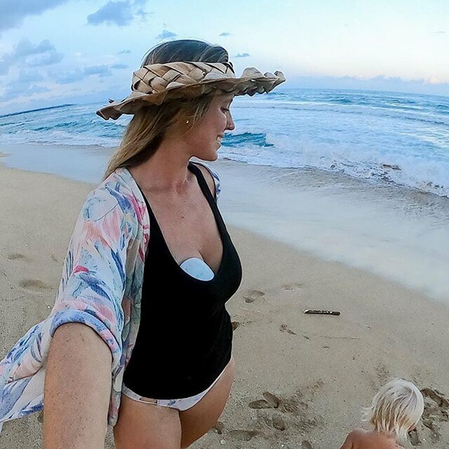 A mother on the beach with an Elvie Pump tucked inside her swimsuit. A blonde child plays in the sand next to her. 