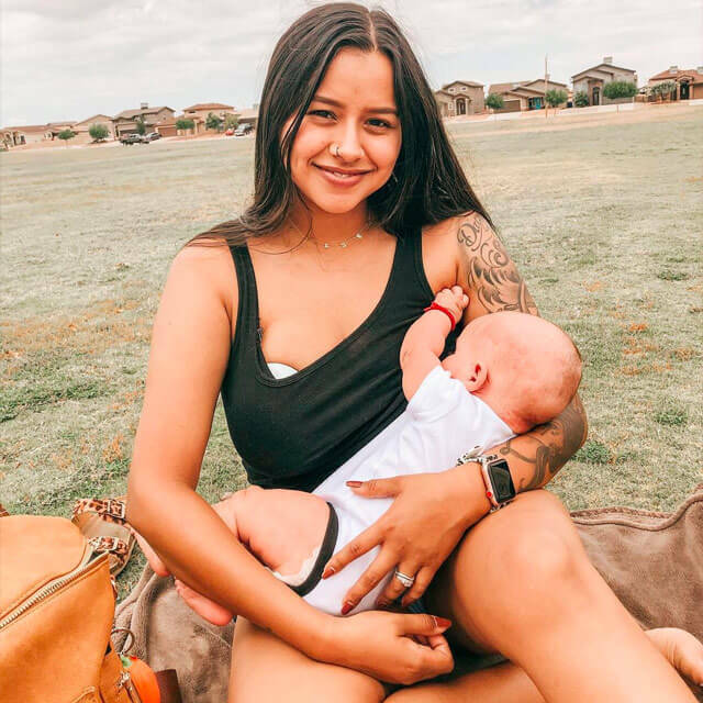 A woman sat down on grass, wearing a black vest top and denim shorts. She is cradling a baby, and an Elvie pump is just visible under her clothes on her right breast