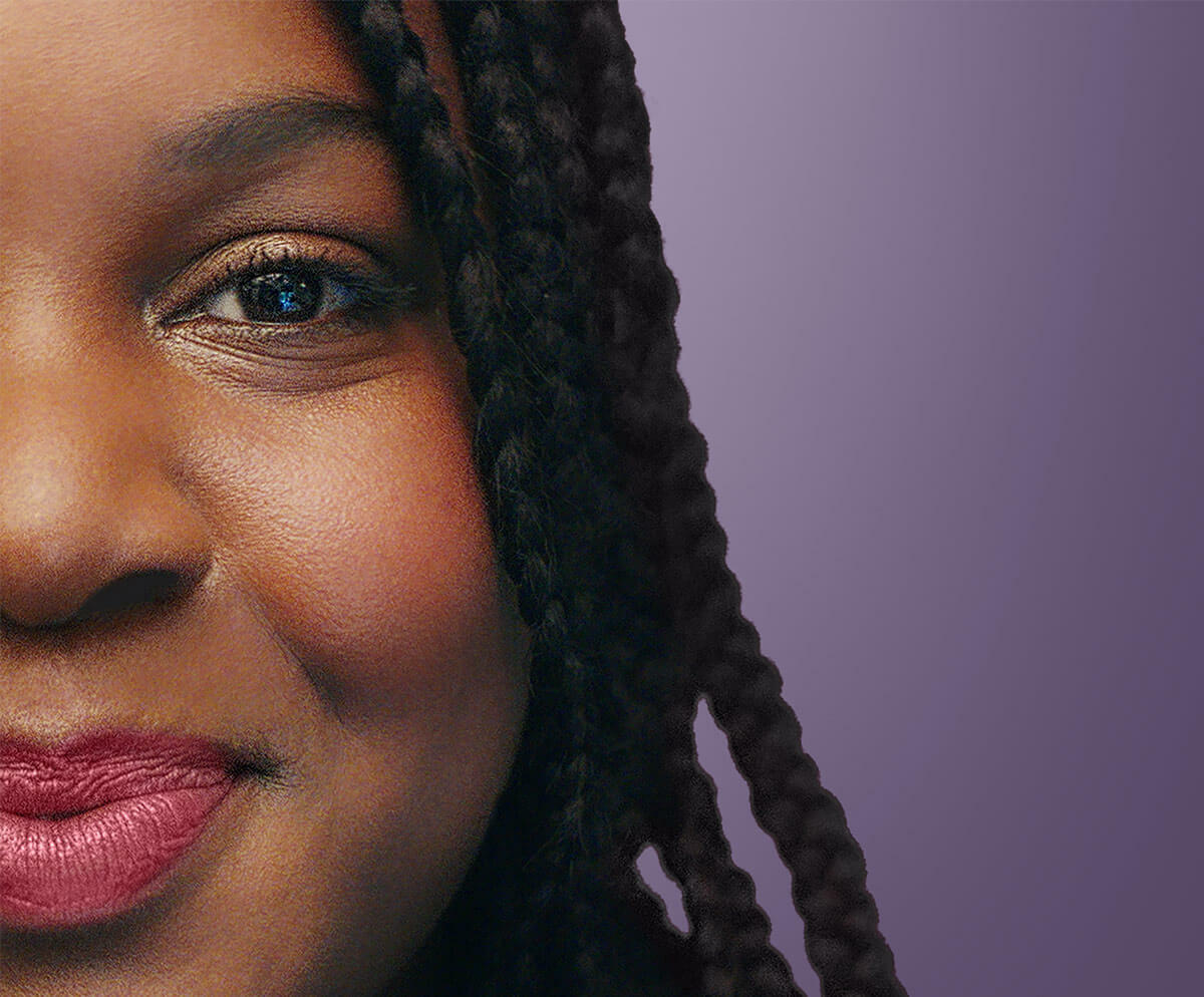 A close up of half a woman's face, smiling and looking at the camera, against a plain purple background