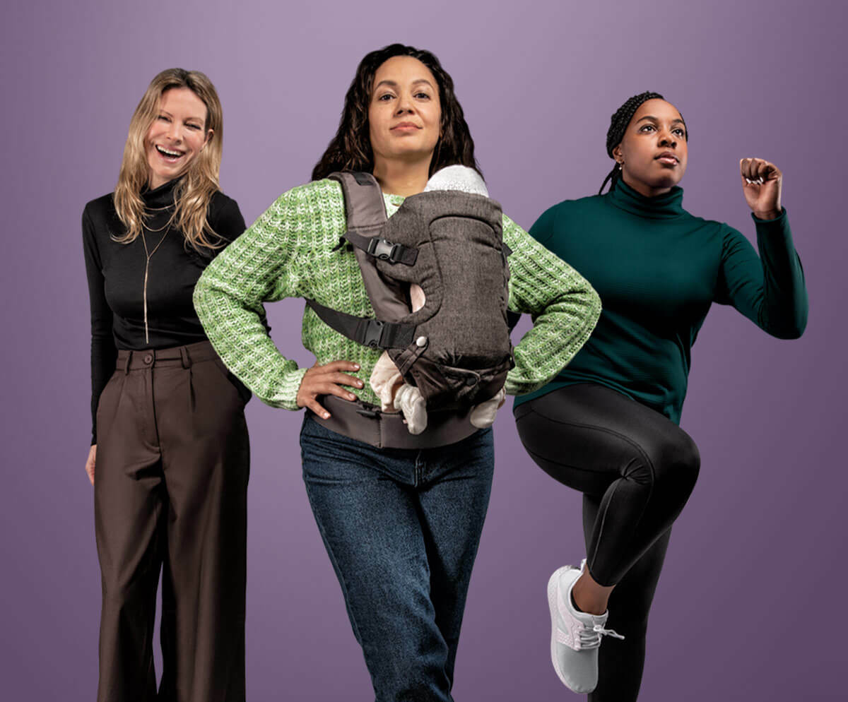 Three women are stood against a plain purple background. The central woman is wearing a green cardigan and has a baby in a carrier on her front. On her left is a woman in smart brown trousers and a black roll neck jumper, and on her right is a woman in black leggings and a teal jumper, her leg raised as if jogging