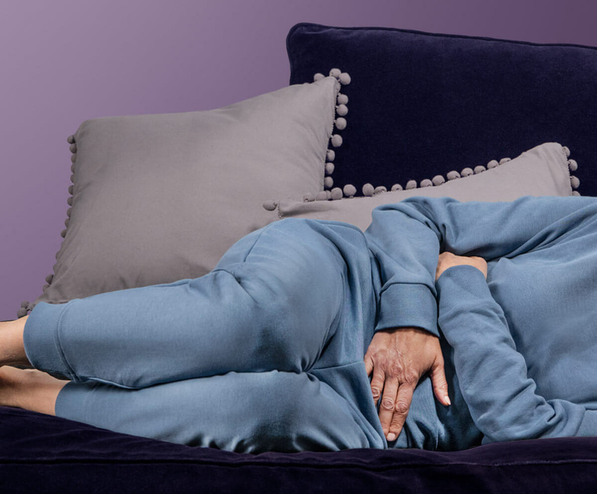 A woman lying on her side on a navy velvet sofa, wearing a blue-grey tracksuit. Her head is not visible in the picture, just her shoulders and the rest of her body. Her legs are bent and one hand rests on her lower abdomen