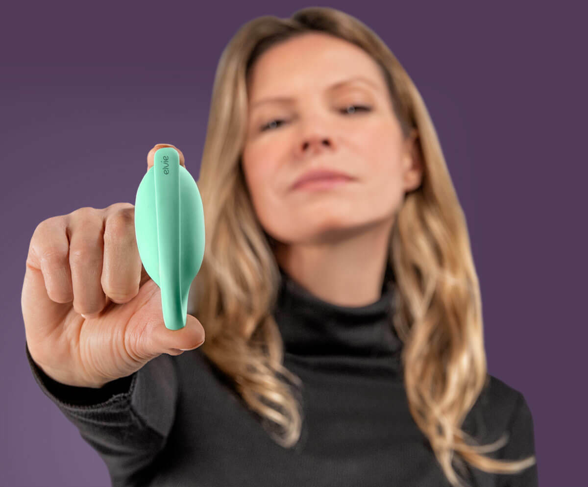 A woman holding an Elvie trainer between her thumb and finger. She is holding it up to the camera and her face is in the background out of focus