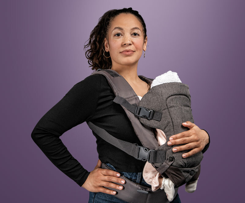 A woman against a plain purple background looking at the camera with a baby in a grey carrier strapped to her front