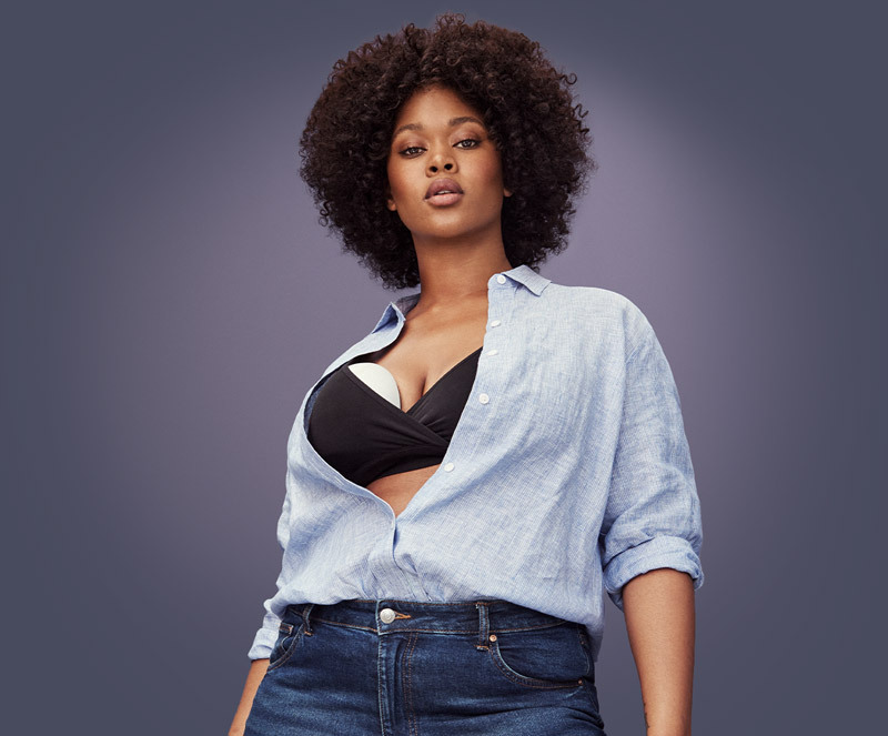 A woman standing looking at the camera with a shirt tucked into jeans, the shirt is unbuttoned enough to see her black bra underneath. There is an Elvie pump visible over the bra on her right breast