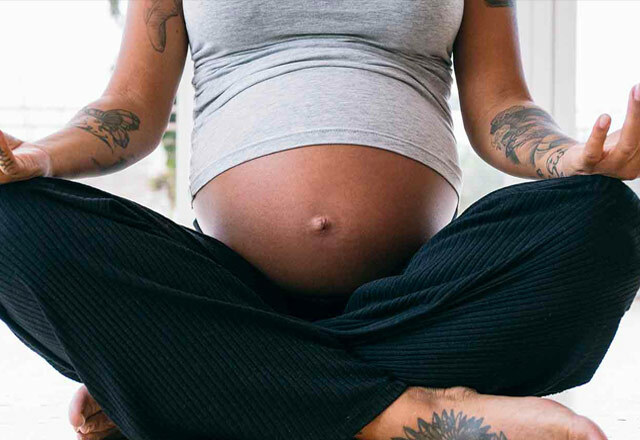 A close up of a woman's legs and pregnant bump. She is wearing loose black trousers and a grey tshirt and sitting cross legged with hands resting on her knees
