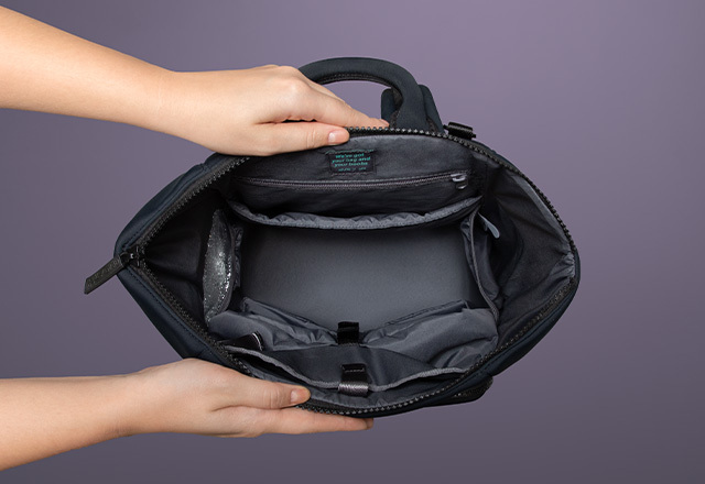 A woman's hands hold the Elvie black rucksack open – it is seen from above as if you're looking straight down into the empty bag. The inside is dark grey with inside pockets and padding