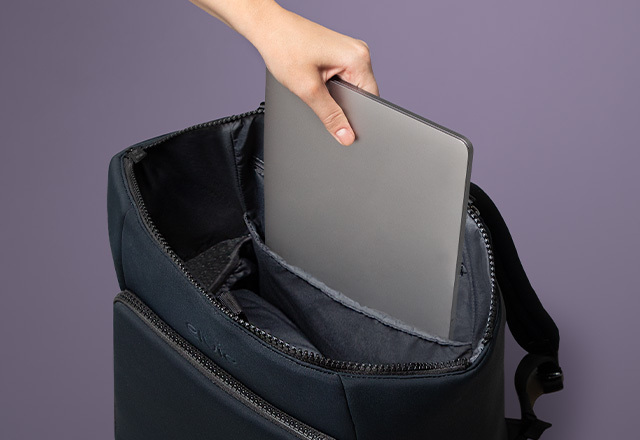 The Elvie black rucksack seen from the top, with the top zip open and an inner pocket visible. A woman's hand is sliding a silver laptop into the inside pocket to demonstrate that it is big enough for a laptop
