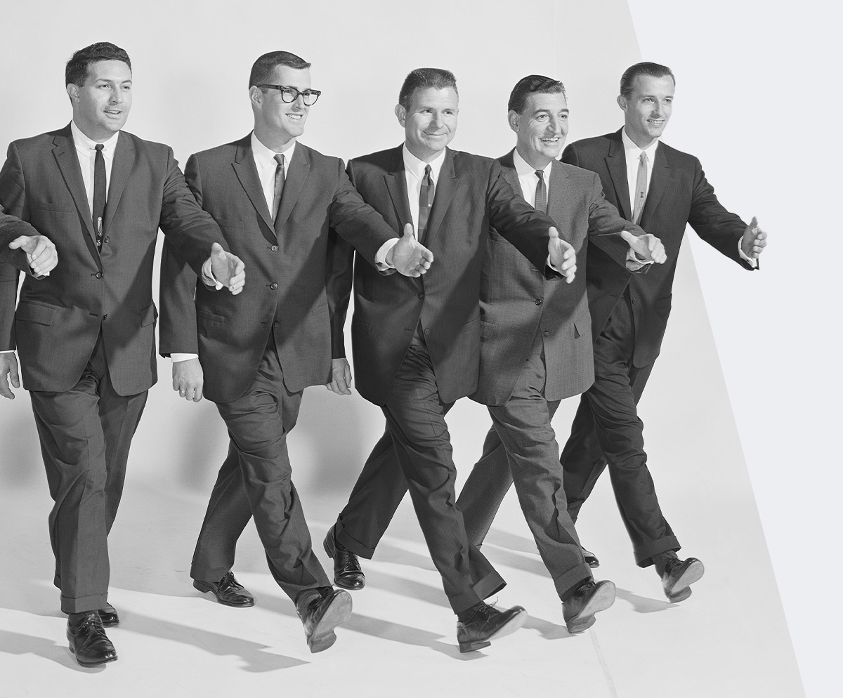 A black and white photo of five smiling men in suits with their arms outstretched, they appear to be walking in unison 