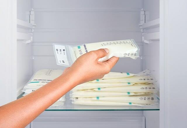 A close up of a fridge shelf with horizontally stacked full milk bags on it. A woman's hand is adding another full milk bag to the stack