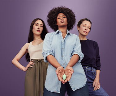 Three women are stood against a plain purple background. The central woman is wearing a green cardigan and has a baby in a carrier on her front. On her left is a woman in smart brown trousers and a black roll neck jumper, and on her right is a woman in black leggings and a teal jumper, her leg raised as if jogging