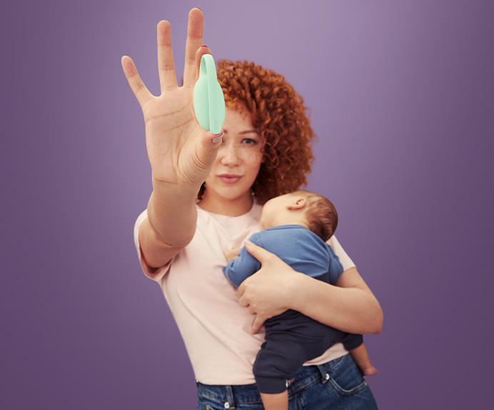 A woman against a plain purple background looking at the camera with a baby in a grey carrier strapped to her front