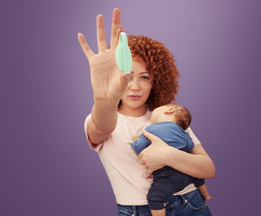 A woman against a plain purple background looking at the camera with a baby in a grey carrier strapped to her front