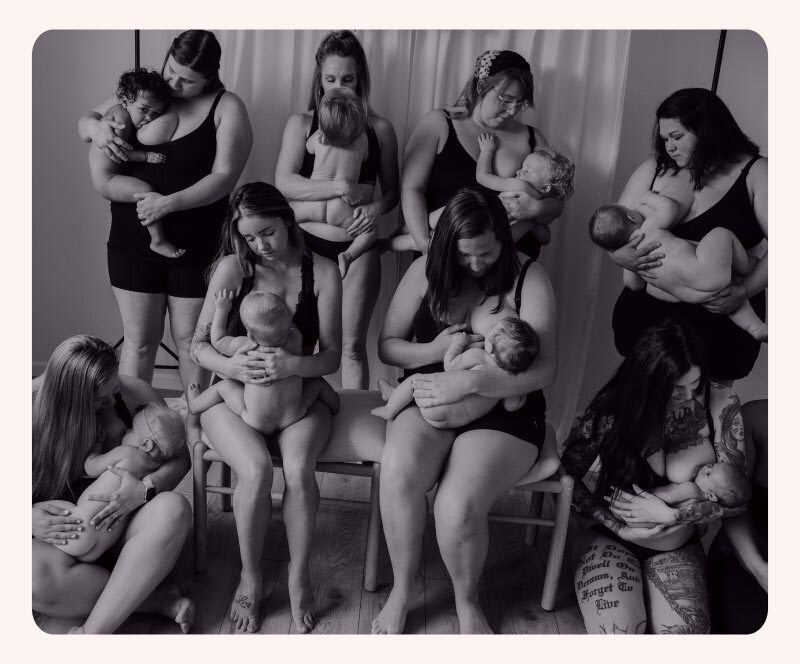 Eight women in their underwear sit on chairs or stand, all breastfeeding their babies. 