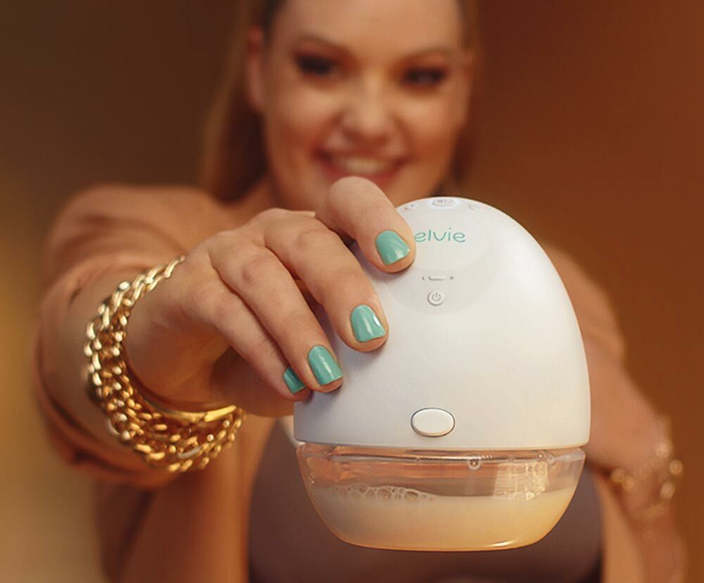 A woman's hand holding an Elvie pump up to the camera, the woman can be seen out of focus in the background