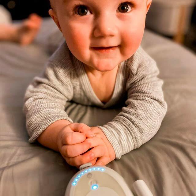 A baby, lying on their front and propping themselves up on their elbows on a grey duvet. In front of them is an Elvie Stride control panel with its lights on