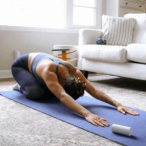 A woman on a yoga mat in a living room, she is in a kneeling position and leaning forwards so her torso is flat against the floor. There is an Elvie Trainer container next to her