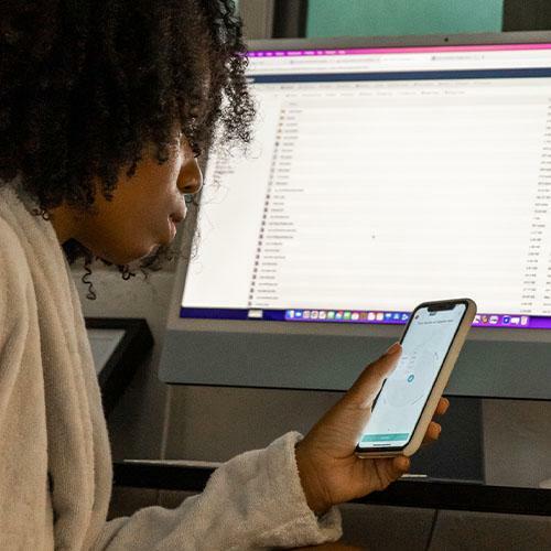 A woman sat at a computer, holding a phone in her hand. She is looking away from the camera and at the phone