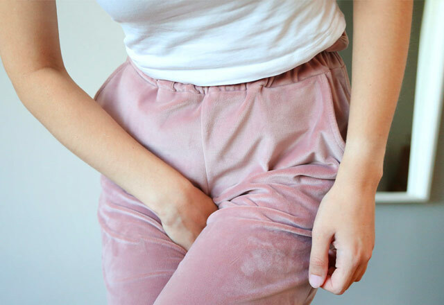 A close up of a woman's lower abdomen and crotch area. She is wearing a white tshirt tucked into pink velour joggers and crossing her legs, her hand clasps at her crotch as if desperate for the toilet