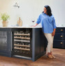 Woman using a black Caple WI6155 Wine Cooler in a modern blue and white kitchen with flowers on disp