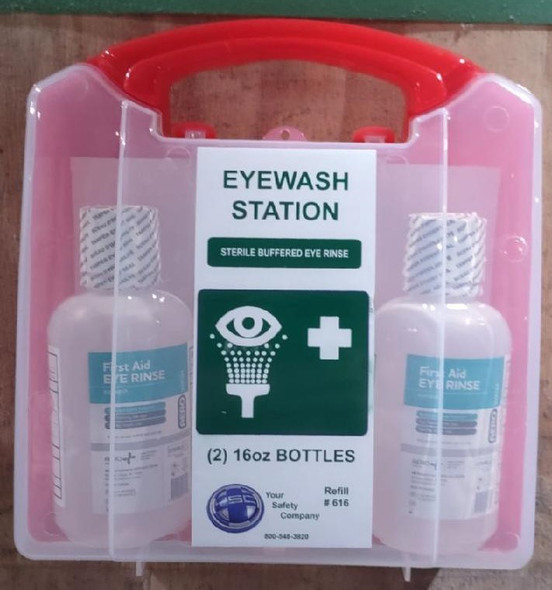 A portable eyewash station in a clear case with a red handle. The kit contains two First Aid Eye Rinse bottles with 16oz of Sterile Buffered Eye Rinse. The central label includes EYEWASH STATION text, a logo, and the Your Safety Company contact information.