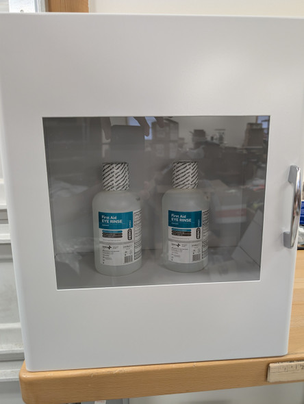 A white, wall-mounted eyewash cabinet is shown, with a clear window on the front. Through the window, two translucent plastic bottles of "First Aid EYE RINSE" are visible, each with a white and gray safety label. The cabinet has a chrome handle on the right side and a white finish, appearing to be brand new and ready for use.