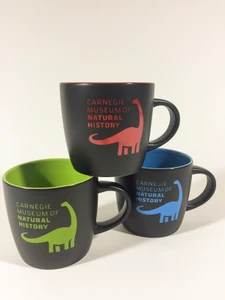 Three stacked mugs in three color variations red, green and blue . Black matte exterior with bright color imprint of dinosaur and museum text logo. Interior of mug is glazed in same color as exterior imprint.