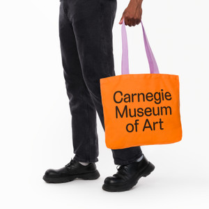 Carnegie Museum of art orange tote with lavender handle, being carried in one hand. Carnegie Museum of art orange tote with lavender handle, being carried in one hand.