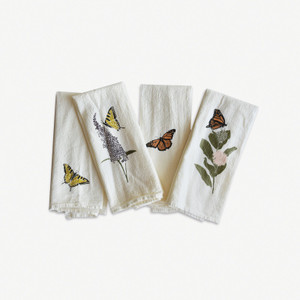 4 off white napkins w/ images of butterflies on them. from left to right: 1. a yellow butterfly on the lower left corner. 2. a yellow butterfly on a purple flower. 3. a monarch in the middle. 4. a monarch above a pink flower