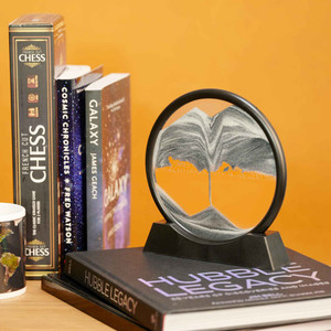 A circular sand art tabletop decoration sitting on top of a coffee table book on a bookshelf.