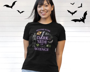 A woman wearing a black t-shirt with the words "Welcom to the Dark Side of Science" surrounded by images of scary science like skeltons, black holes, and poison