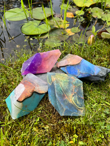 Image shows example of the four SoapRock varieties next to a pond. At the top is Amethyst, a purple and pink soap. Clockwise from that are the Black Opal, a blue and pink soap with gold veins, followed by Labradorite, a multifaceted light blue soap with gold veins, and finally with  Turquoise, a turquoise blue and pink soap with a variety of lighter tones.