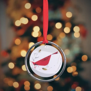 This circular double-sided metal ornament features the 1954 "Cardinal with Berries" illustration of a  bright red cardinal perched among angular, stark branches while consuming vibrant clusters of berries in the "minimal realism" and geometric style Charley Harper was known for. A silver ring hangs separately, with a red ribbon attached to this for hanging.
