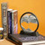 A circular sand art tabletop decoration sitting on top of a coffee table book on a bookshelf.