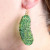 Green pickle shaped acetate earrings on a golden hook shown on a persons ear.