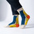 Abstract socks featuring line drawn faces and the colors yellow, blue, and orange -- Photographed worn on feet, styled with dark skinny jeans.