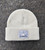 A photo of the bone beanie. The knit texture is visible. Centered on the cuff is a silver patch bearing the CMNH Logo. This includes text reading "CMNH" and "Established 1895" above and flanking the silhouette of Dippy, the CMNH logo, in bone. "Carnegie Museum of Natural History" is written below this.