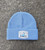 A photo of the powder blue beanie. The knit texture is visible. Centered on the cuff is a silver patch bearing the CMNH Logo. This includes text reading "CMNH" and "Established 1895" above and flanking the silhouette of Dippy, the CMNH logo, in light blue. "Carnegie Museum of Natural History" is written below this.