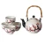 A photograph of the tea set. The tea pot is a traditional shape, with a top lid and detachable bamboo handle. It has a white base, black spout, and black accents on the ball atop its lid. Both the tea pot and tea cups are white, decorated with a cherry blossom branch -- the branch is black, with red and pink blossoms.