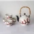 A photograph of the tea set. The tea pot is a traditional shape, with a top lid and detachable bamboo handle. It has a white base, black spout, and black accents on the ball atop its lid. Both the tea pot and tea cups are white, decorated with a cherry blossom branch -- the branch is black, with red and pink blossoms.