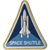 This image shows a white space shuttle  on a 2 tone blue background.  It is triangular and is outlined in gold thread.