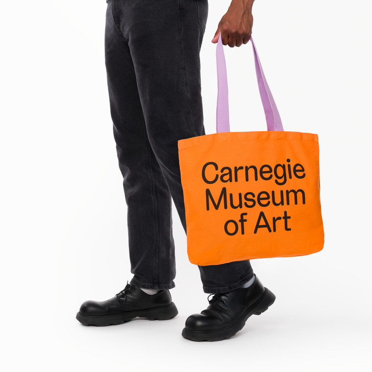 Carnegie Museum of art orange tote with lavender handle, being carried in one hand.