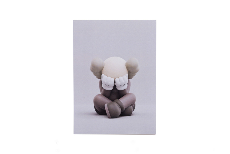 KAWS companion figure sitting cross-legged, hands on face on a postcard.