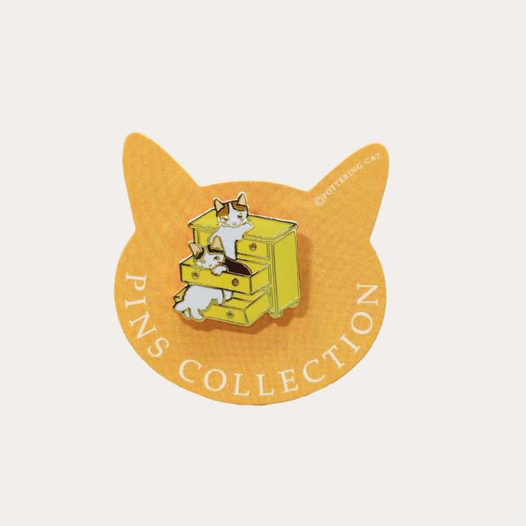 Cat head shaped paper backing card, enamel pin of cats playing in drawers of a dresser.