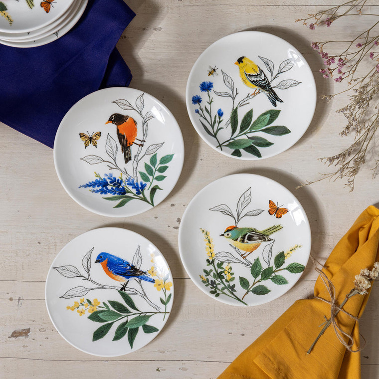 four white plates with different colored birds printed on them on a table with napkins and flowers