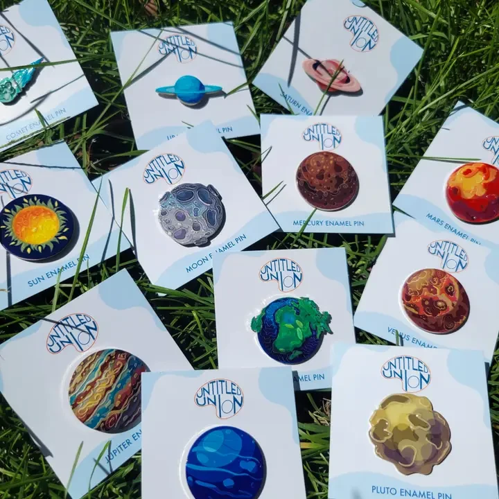 An assortment of enamel pins featuring illustrations of the Sun, eight planets, Pluto, and a comet