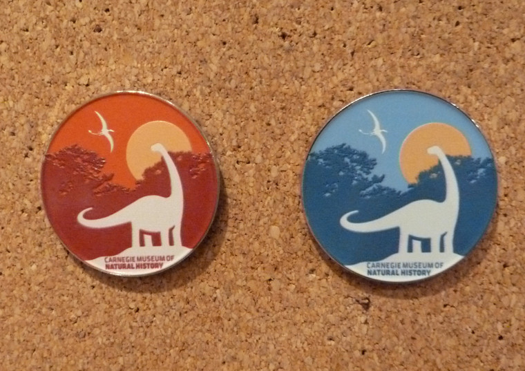 Image shows both pin variants side by side on a cork board. They are circular, 1.5" in diameter, and feature a white silhouette of Dippy the Diplodocus in front of a forest alongside a smaller pterodactyl.  A circular sun is visible in the right behind Dippy in yellow. Below Dippy the text "Carnegie Museum of Natural History" is visible in white. The pins have a silver border.